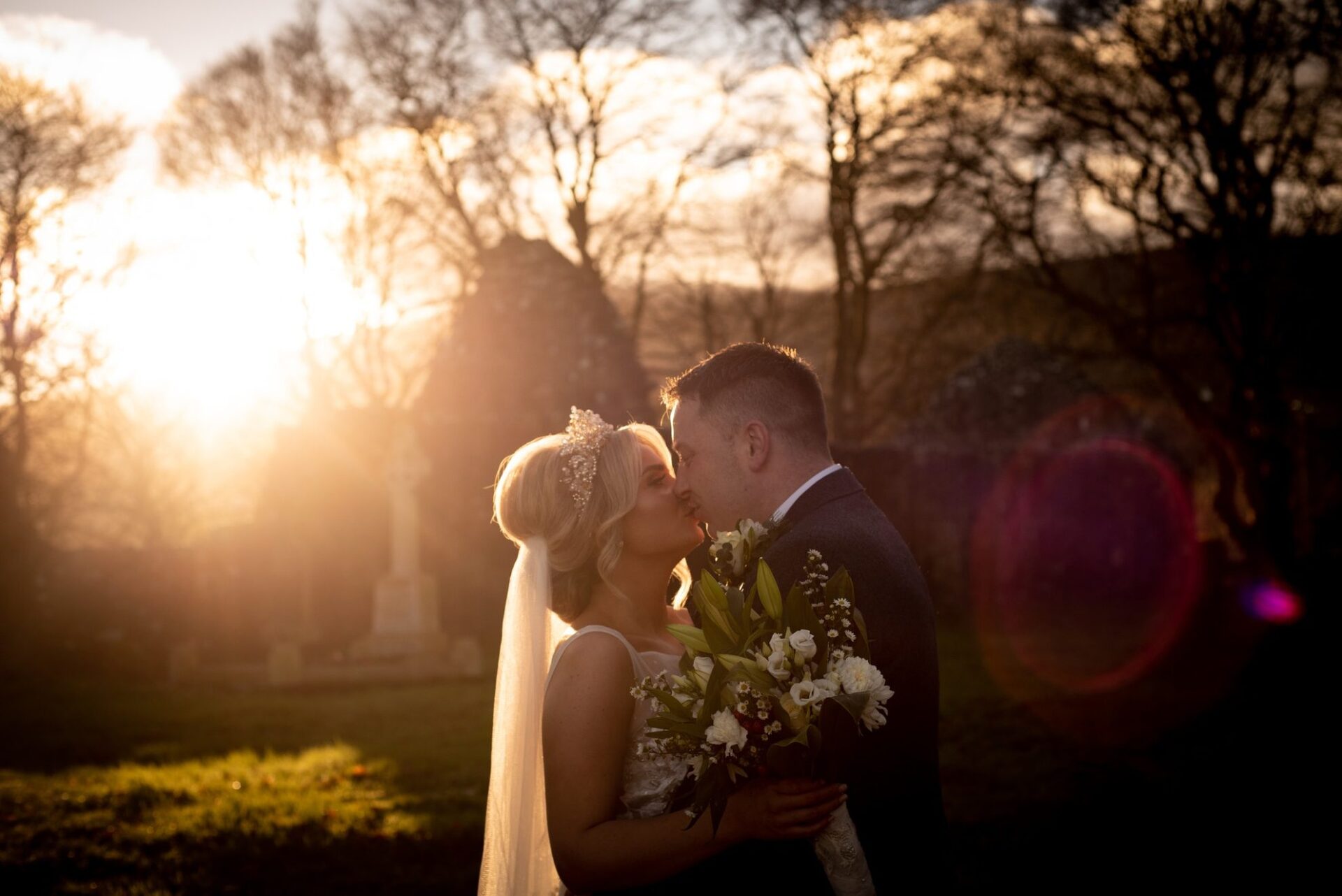 A bride and groom kissing as the sun sets between the trees.