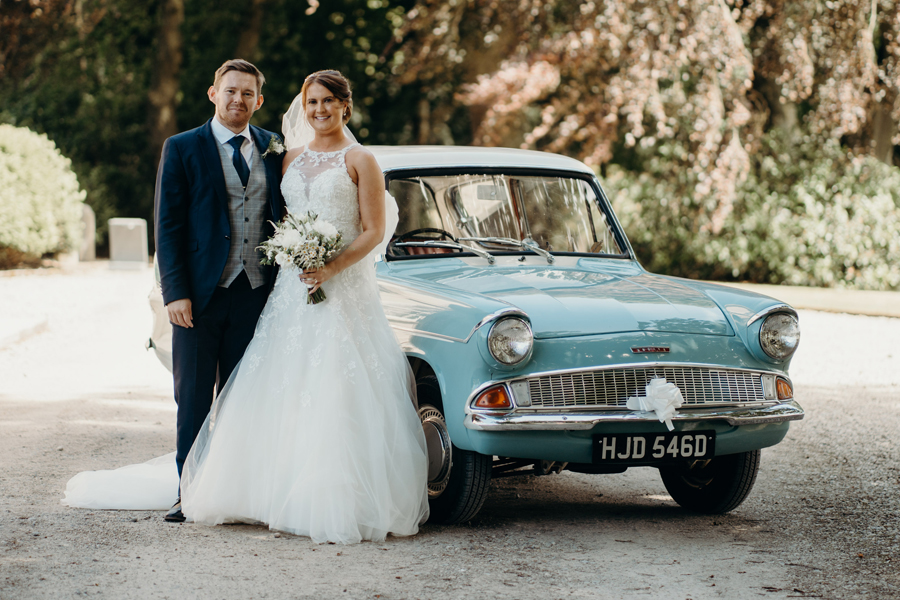 A bride and groom stand next to a blue 1965 Ford Anglia, a replica of the car that appeared in Harry Potter.