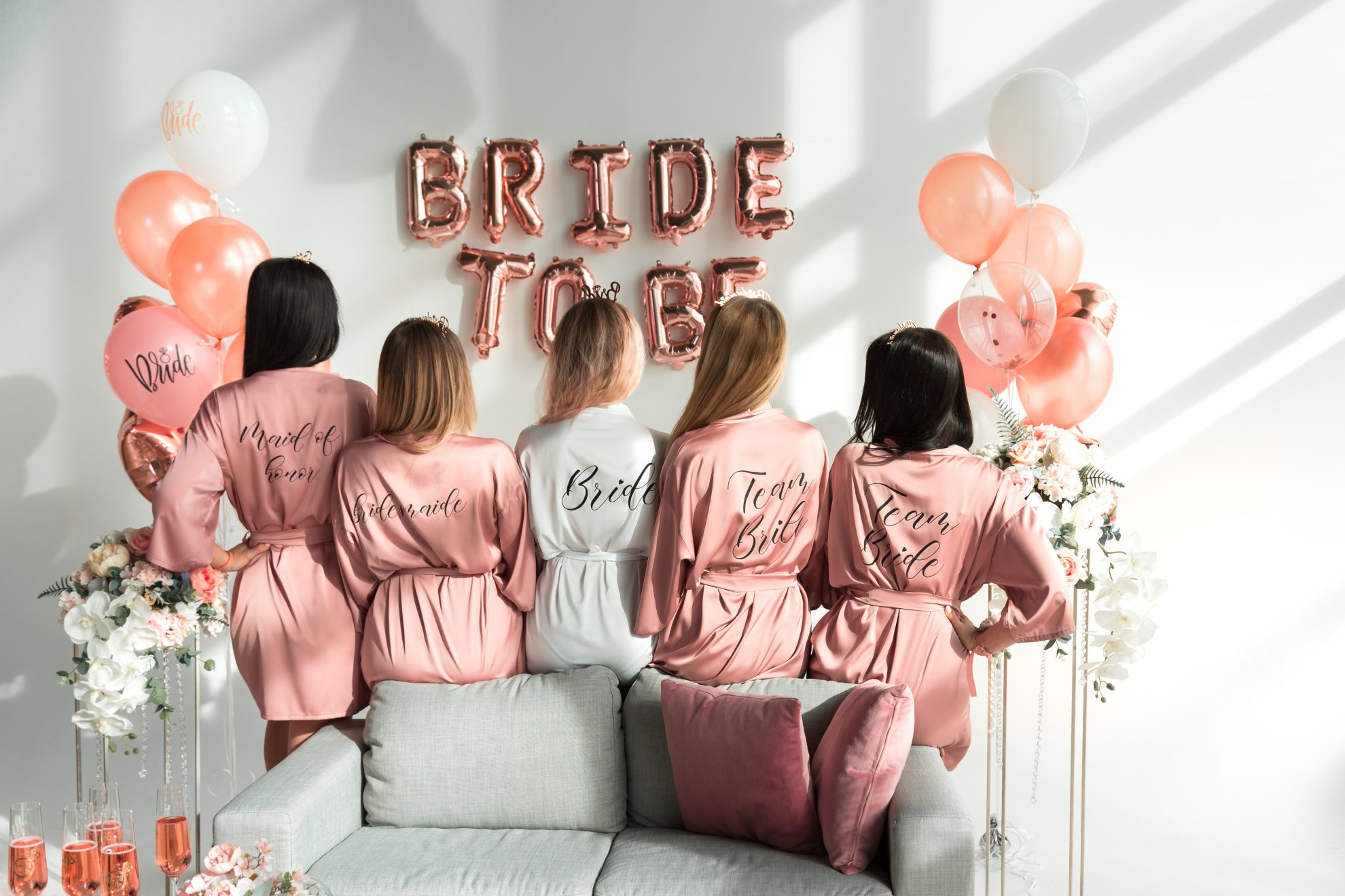 A bride stands in the middle of her 4 bridesmaids. All the ladies in the picture are facing away from the camera and the robes they are wearing either states 'bride' 'Maid of Honour' 'Bridesmaid' or 'Bride Tribe.' Balloons can be been in the background which spell 'Bride to be'