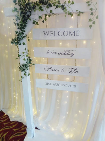 This is a beautiful rustic welcome wedding sign that is paired with green foliage and set against a fairy light backdrop.