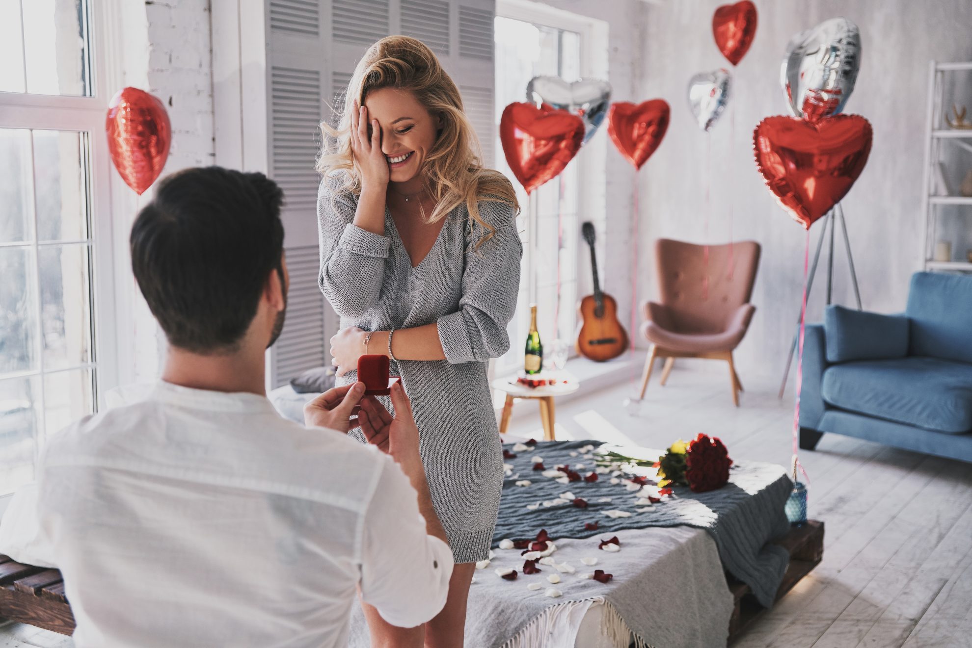 A woman looks happy as a male is on one knee in front holding an engagement ring box. Red and silver heart balloons are seen in the background. Image used in the 8 signs your partner is going to propose article.