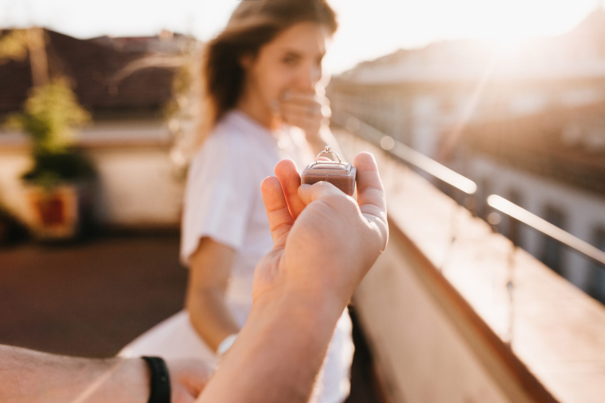 A male hand holds an engagaement ring. We can see a surprised looking woman in the background. Image used in the 8 signs your partner is going to propose article.