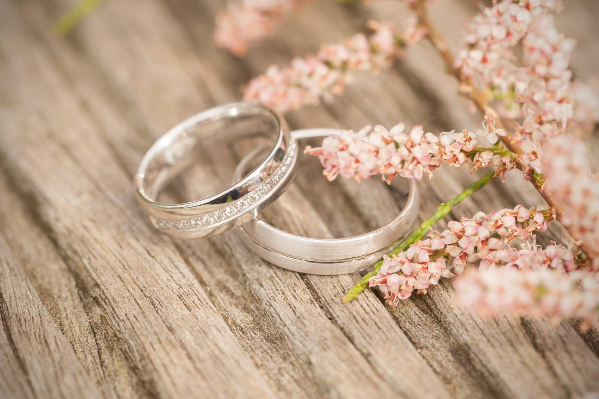 Two wedding rings sit on a table with pink flowers beside them.