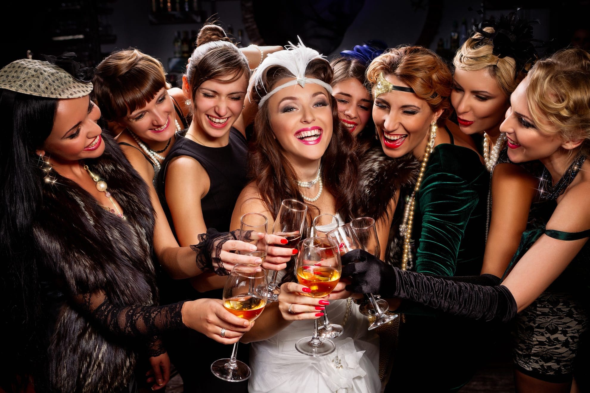 A bride smiles while a group of friends surround her. They are clinking glasses and are wearing fancy dress. Image used in the 8 Hen Party Ideas for Every Type of Bride article.