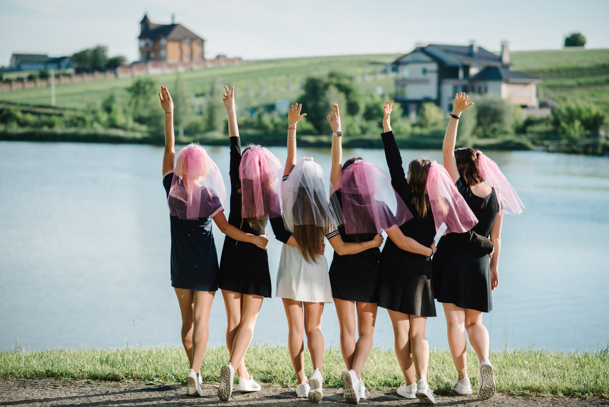 A bride and her hens pose in front of a lake. The bride is in a while outfit with a white veil while the others are in black outfits with pink veil. Image used in the 8 Hen Party Ideas for Every Type of Bride article.