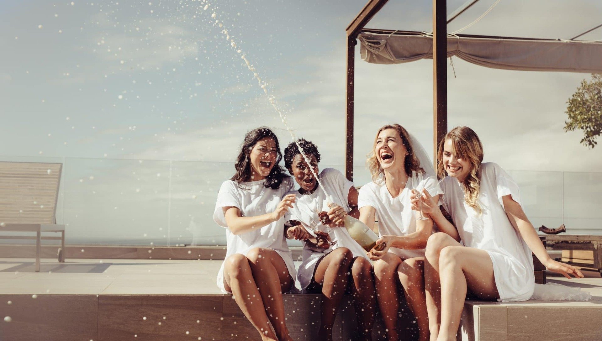 Four ladies sit outside and one pops a bottle of champagne.